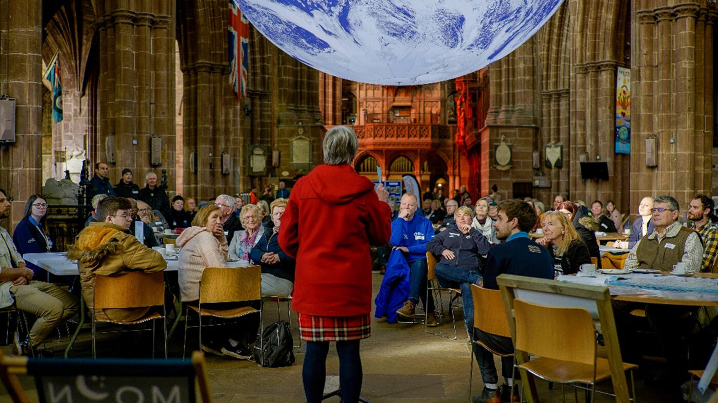 A person in a red coat gives a talk to an audience of people sat at tables as part of Chester Zoo's Wildlife Champions programme. The talk is taking place in Chester Cathedral, and suspended from the ceiling is a large globe.