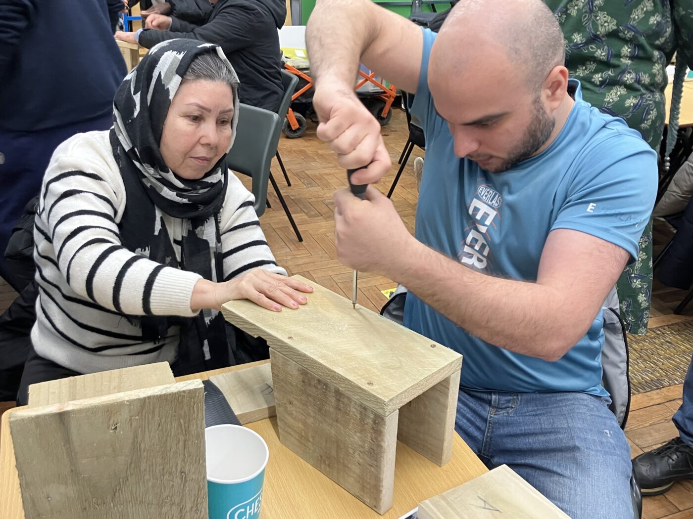 A human female wearing a black head scarf and a black and white stripey top hold a wooden bird box on a table whilst a human male wearing a blue tshirt usese a screw driver to fix the box together using screws.