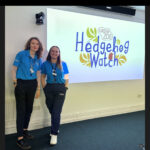 Two members of staff in Chester Zoo uniform stand in front of a Hedgehog Watch sign