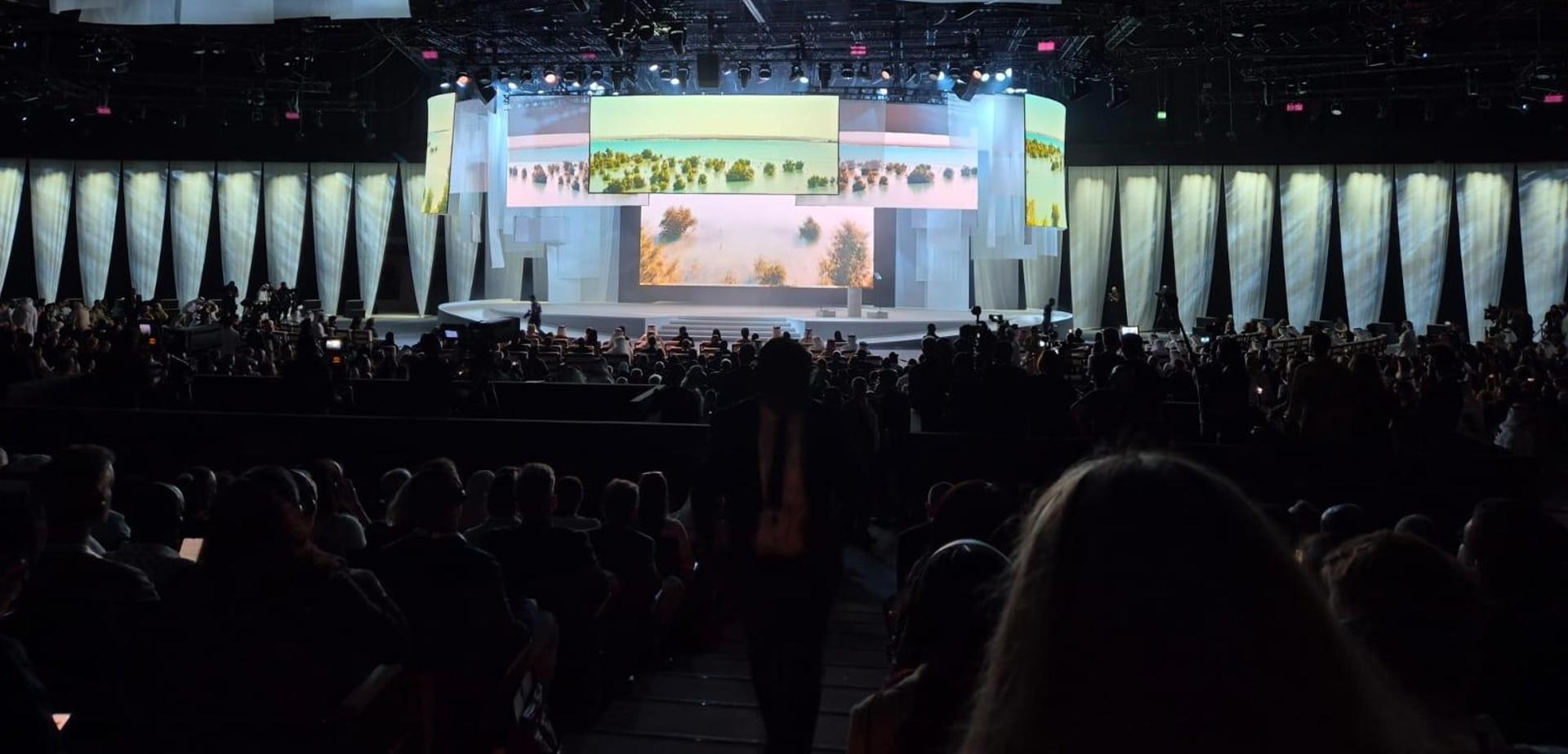 A crowded conference hall. It's dark, except for the brightly lit stage covered in screens showing nature scenes