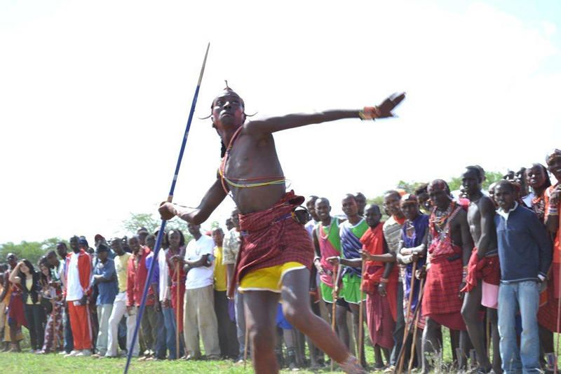 A Maasai moran (warrior) poised to fling a javelin. There is a line of spectators behind him