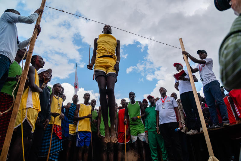 A maasai warrior in yellow colours jumping so his head touches a string marker. He is surrounded by spectators
