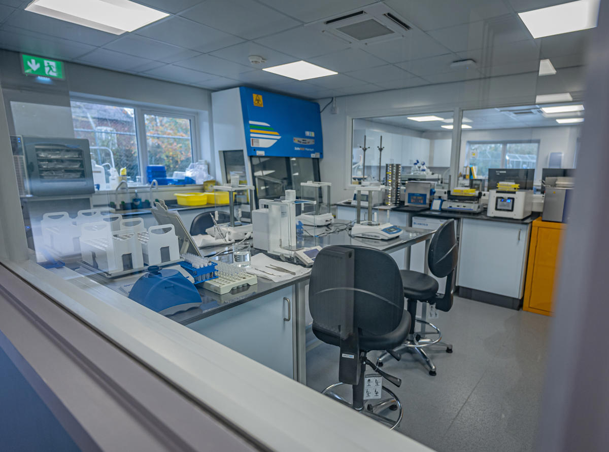 Image shows a lab viewed through a glass window. The lab is kitted out with racks of test tubes and other equipment