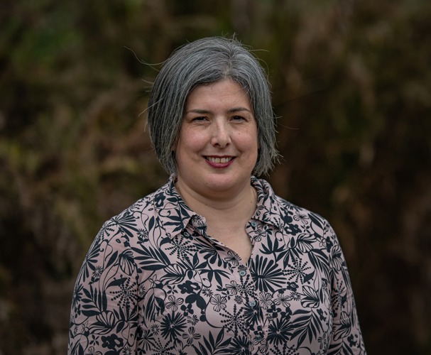 Charlotte Smith - a woman with chin-length grey hair and faintly freckled skin. She is smiling at the camera and is wearing a floral black and pink shirt