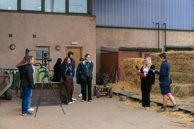 Photo shows a group of young people video and audio recording speakers near bales of straw