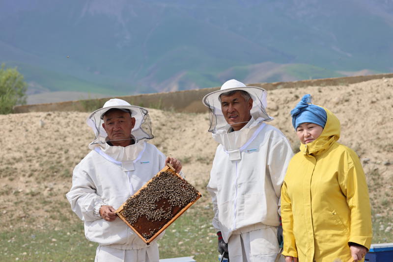 Image shows people in a mountain environment in beekeeping suits. One of them is holding a frame of honeycomb.