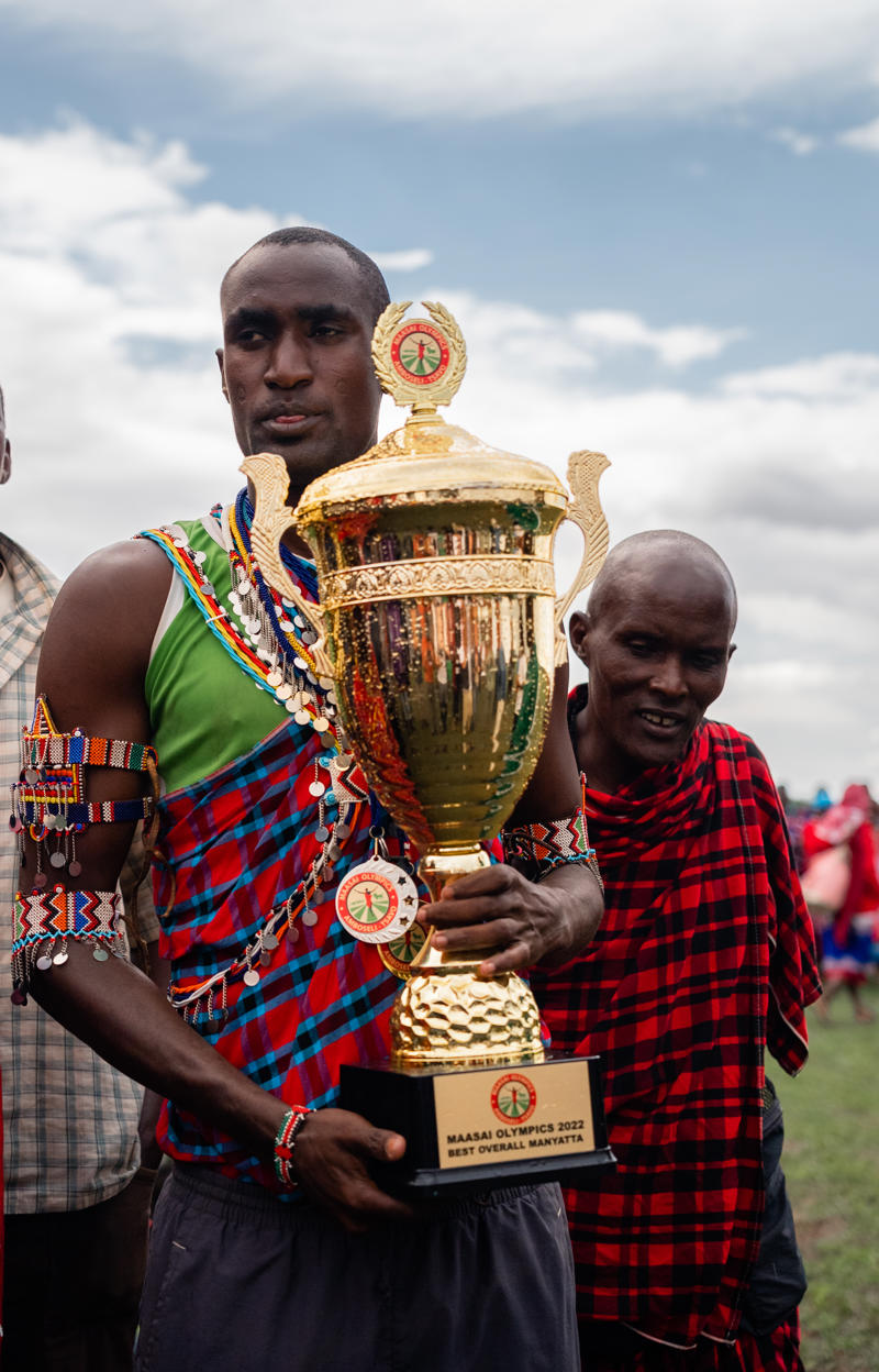 A Maasai man holding a golden trophy. He is wearing traditional ornamentation and clothing.